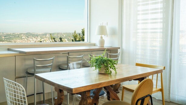 A dining area with a wooden table, chairs, a potted plant, and a view of a cityscape through a window. - Villa Celestina (Marco Simone)