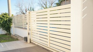 A white metal gate with horizontal slats, a concrete pathway, and a small trash bin. - Villa Celestina (Marco Simone)