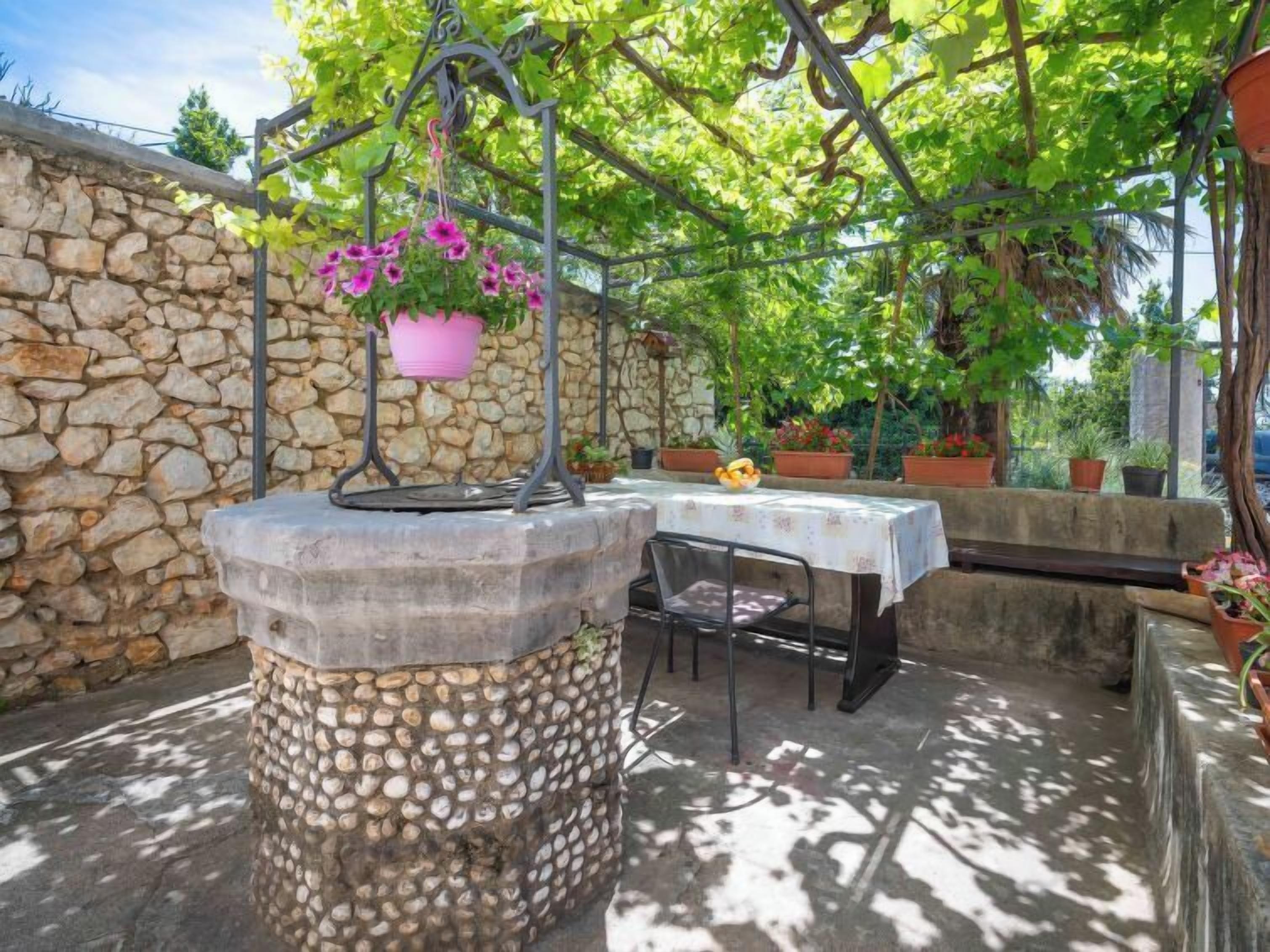 A stone wall with a hanging planter of pink flowers, a table with a white tablecloth, and a bench.