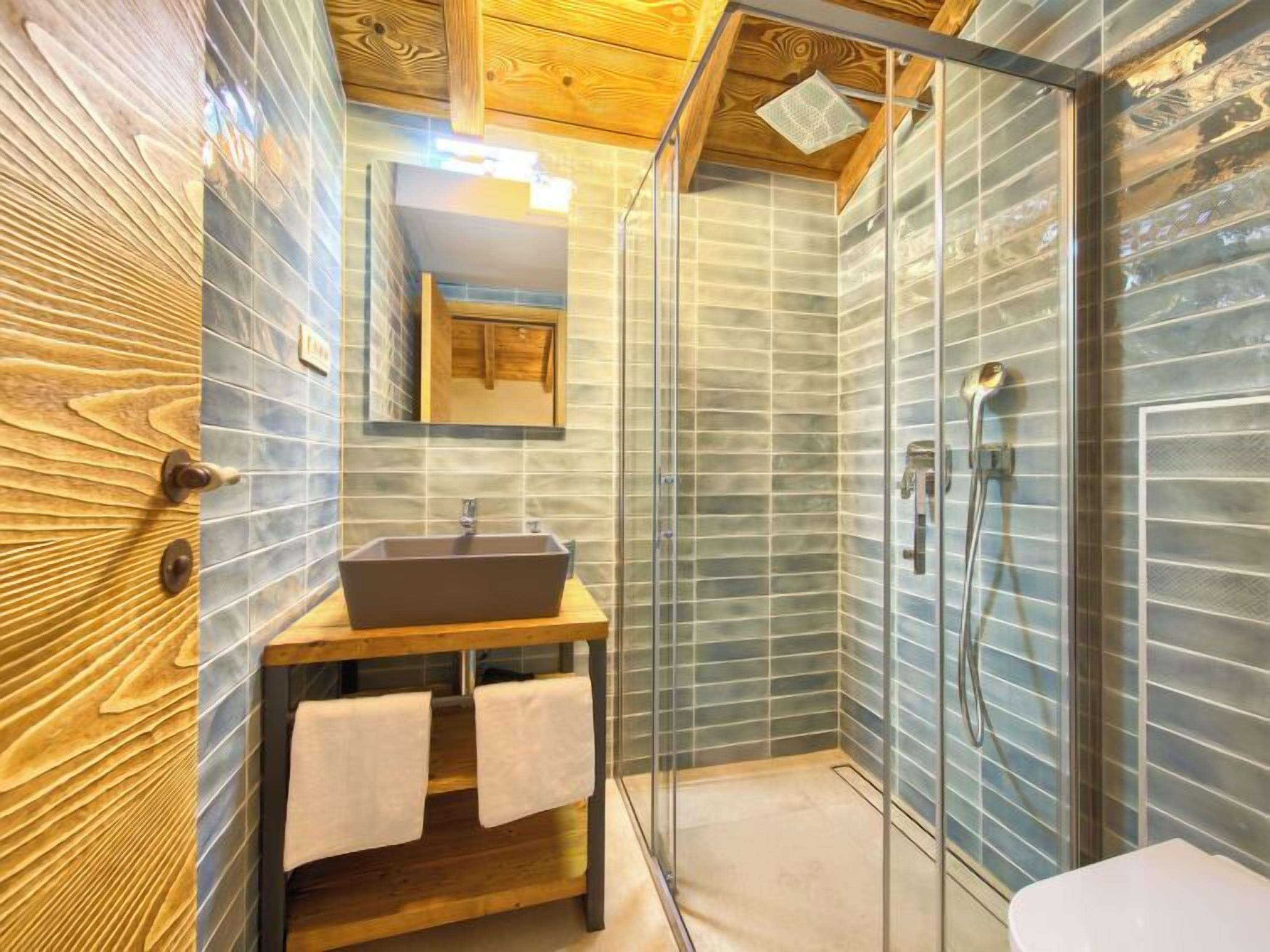 A modern bathroom with a wooden vanity, a large mirror, a glass shower enclosure, and a tiled wall.