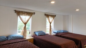 Une chambre avec deux lits simples, chacun agrémenté d’un oreiller bleu et d’une couverture marron. Il y a des fenêtres avec des rideaux beiges, et une petite table entre les lits.