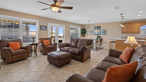 A spacious living room with leather sofas and ottomans, a ceiling fan, and a kitchen area in the background.