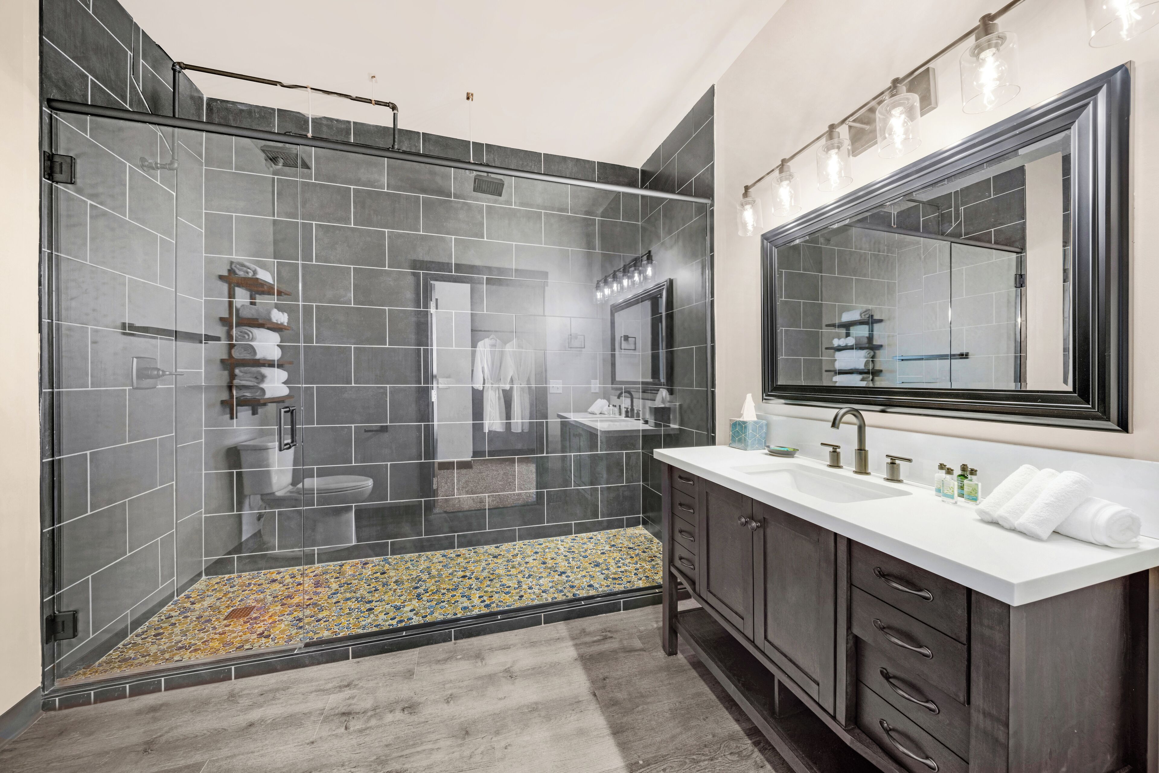 Un baño moderno con ducha envuelta en vidrio, paredes de azulejos oscuros, un lavamanos blanco y un espejo grande.