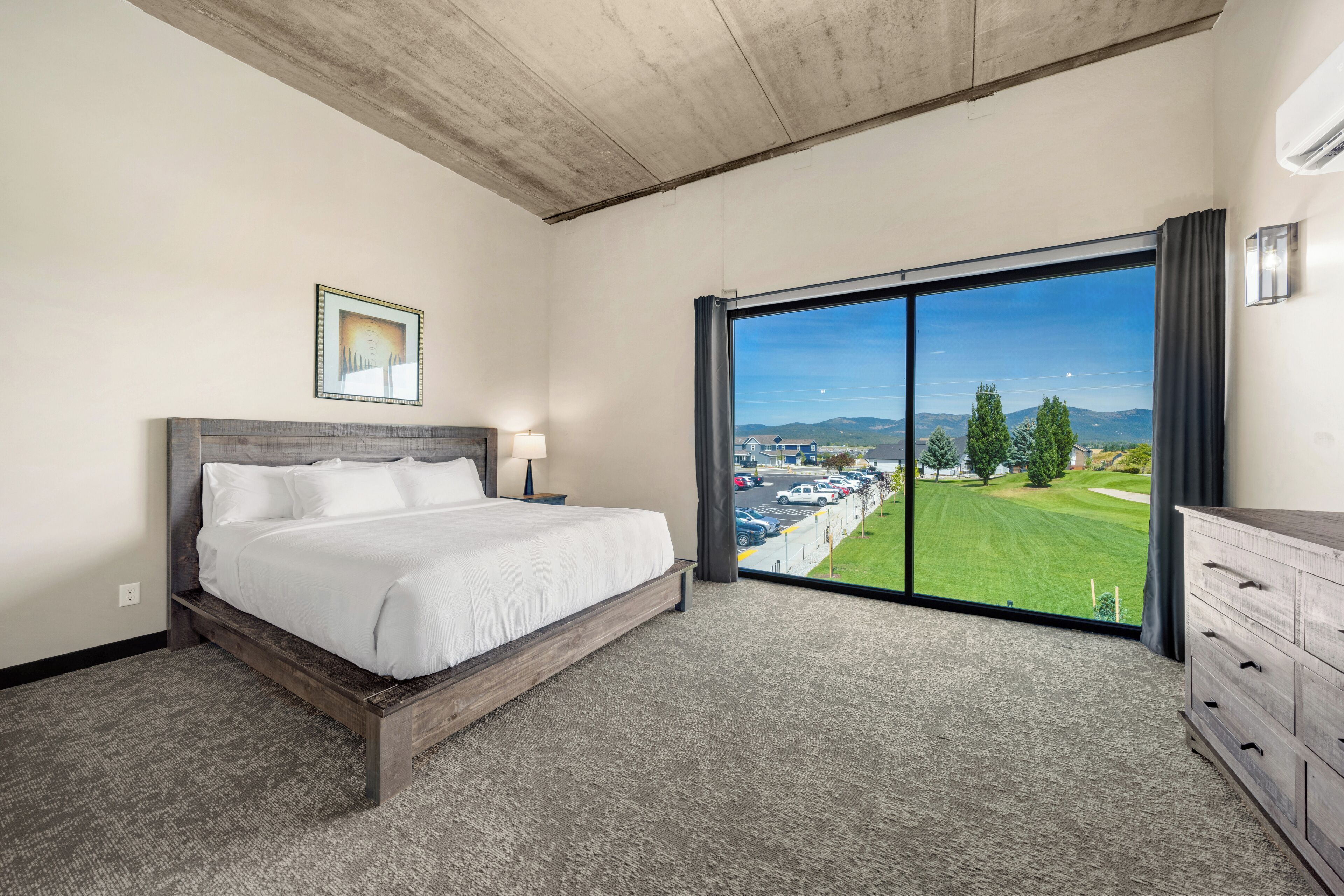 Ein Schlafzimmer mit einem großen Bett, einem Nachttisch, einer Kommode und Blick auf eine grüne Landschaft durch ein großes Fenster.