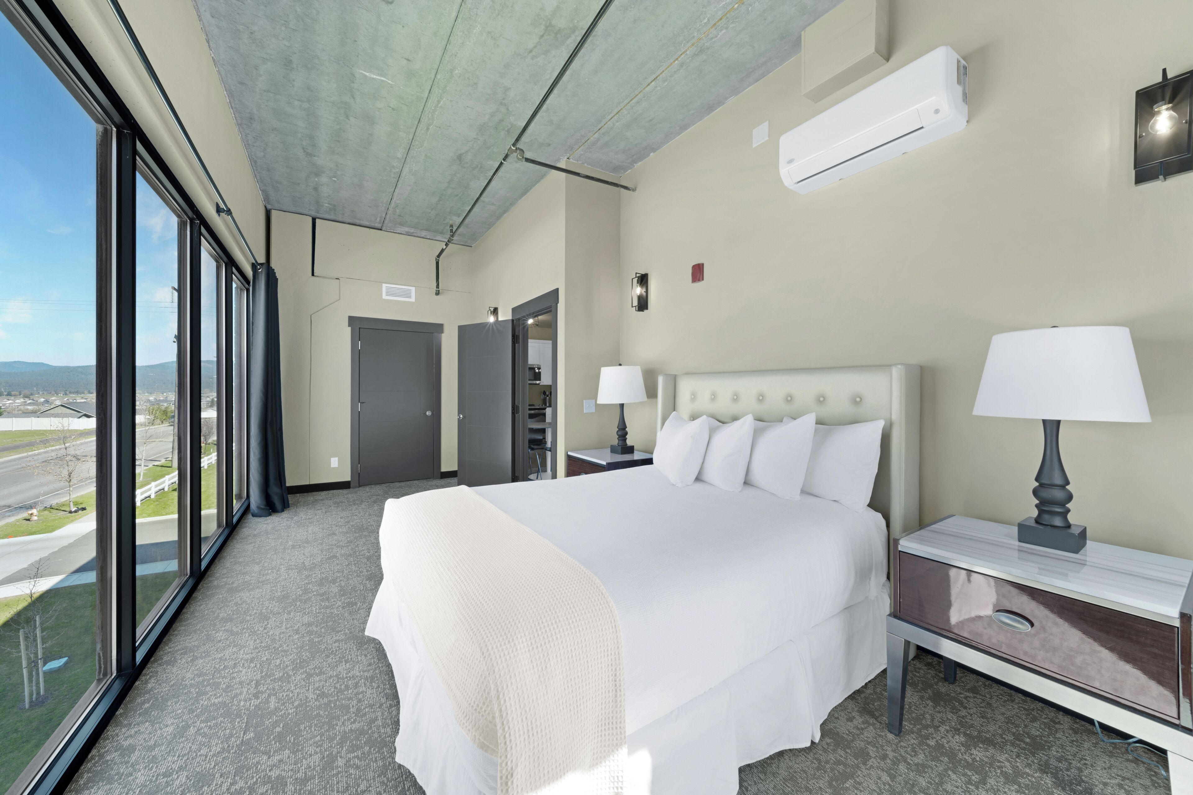 Hotellihuone, jossa on suuri sänky, kaksi yöpöytää valaisimineen, näkymä ulos suurista ikkunoista ja ilmastointilaite seinällä.