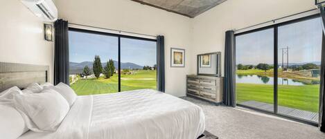 Un dormitorio con una cama grande, un aparador y un televisor. Tiene ventanas amplias con vistas a un campo de golf y un lago.