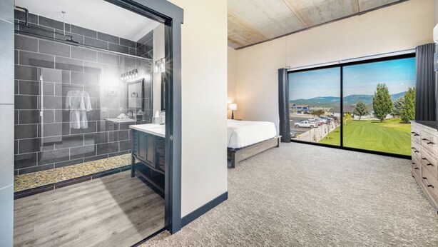 A modern hotel room with a shower, a bed, a large window with a view of the parking lot and hills, and a wooden cabinet. - The Suites at Prairie Falls & Event Center (Post Falls)