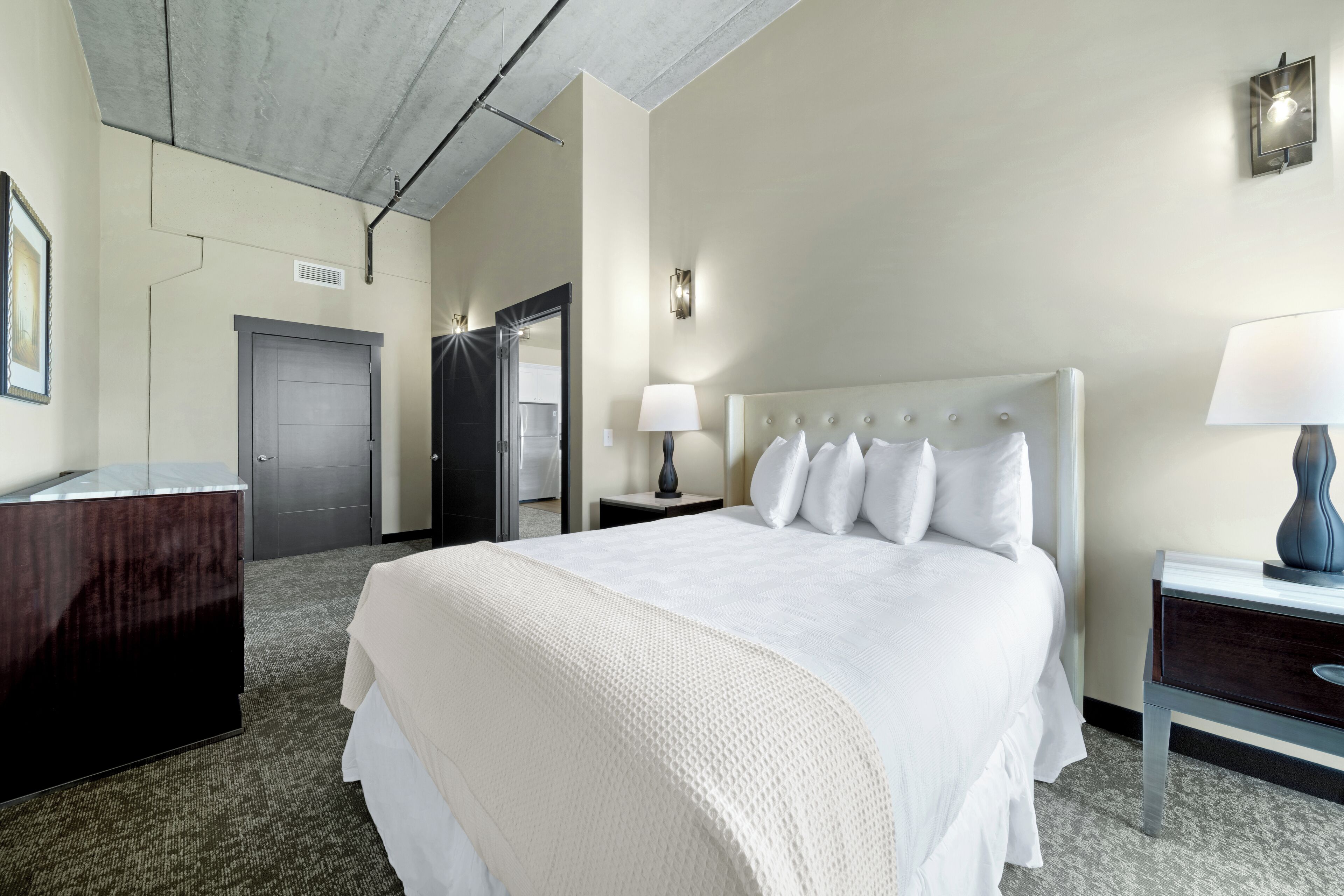 A hotel room with a large bed, a bedside table with a lamp, a wardrobe, and a door leading to another room.