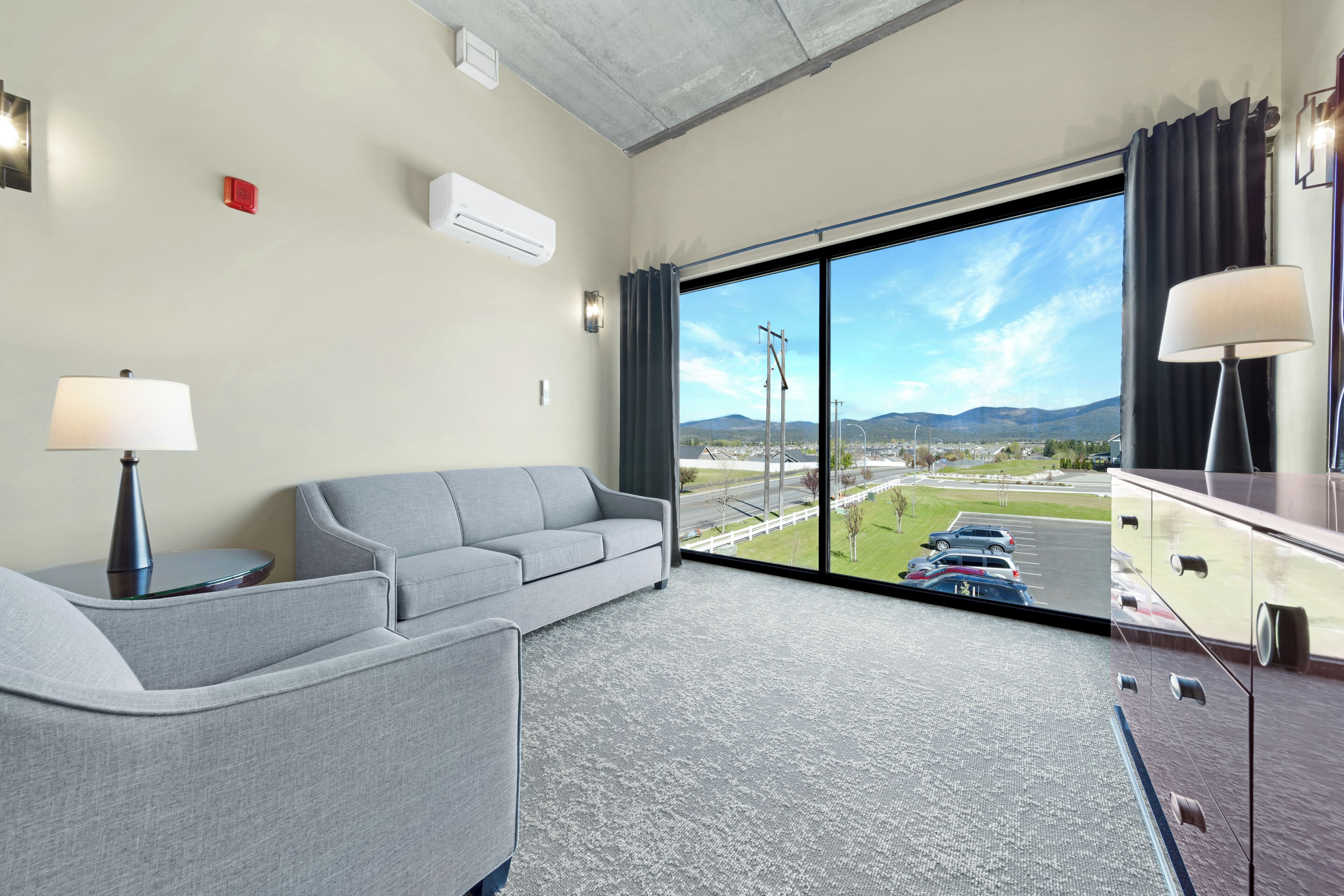 A hotel room with a large window, a grey sofa, a side table with a lamp, and a view of a parking lot and mountains.