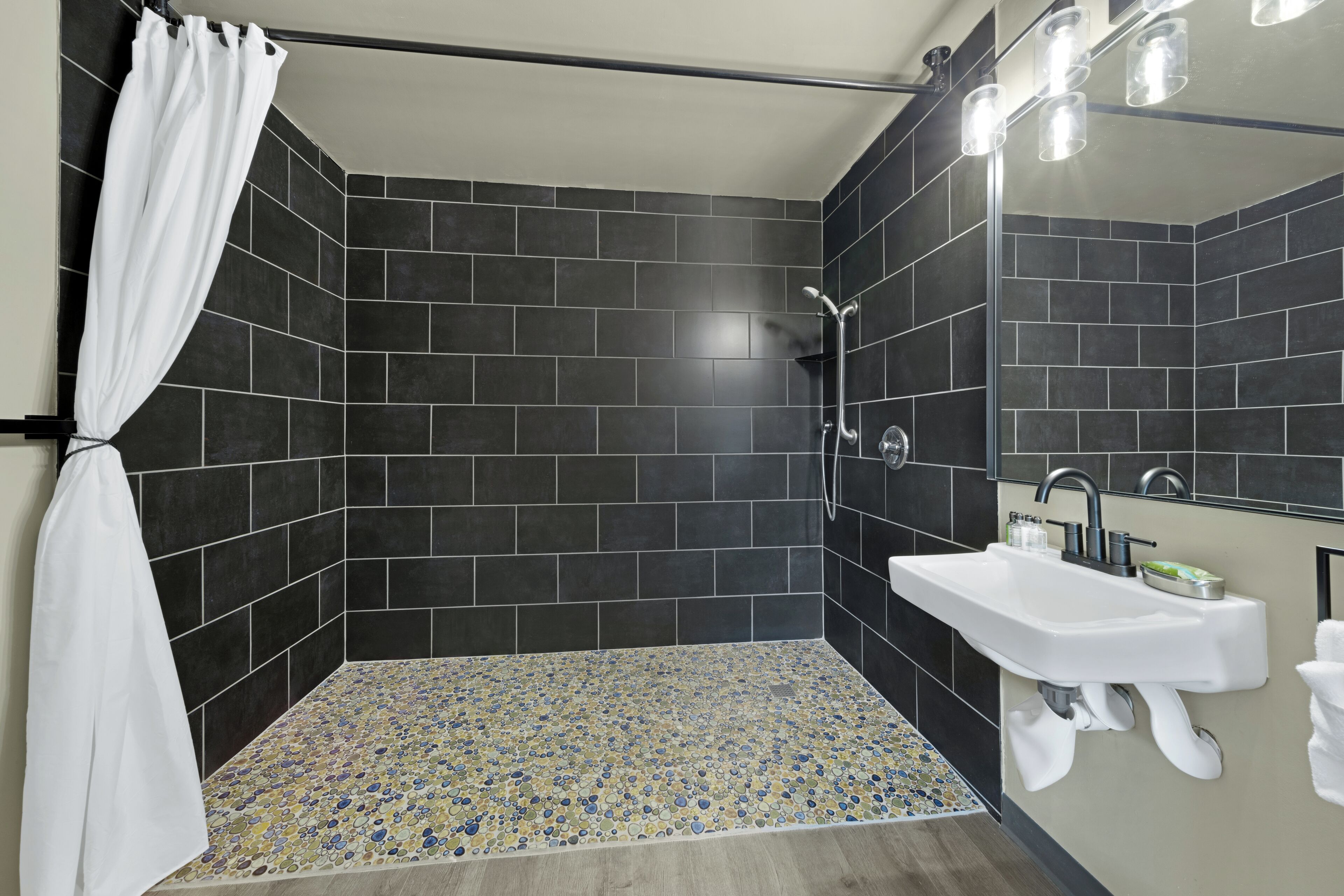 Un bany modern amb rajoles fosques, una dutxa amb porta de vidre, un lavabo amb mirall i un paviment amb dibuixos.