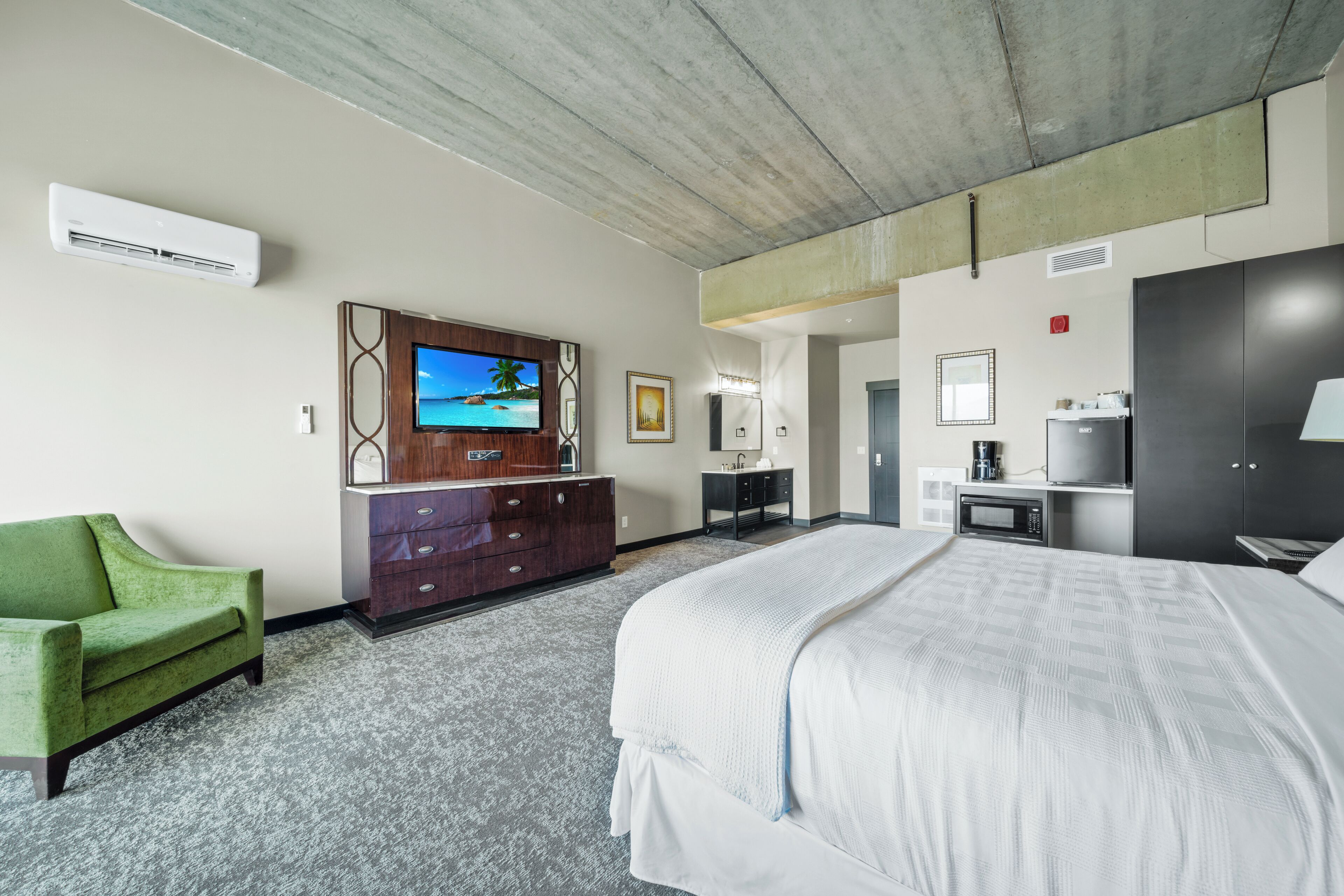 Ein Hotelzimmer mit einem großen Bett, einem grünen Sessel, einem Fernseher, einem Schminktisch, einer Mikrowelle und einem Kleiderschrank.