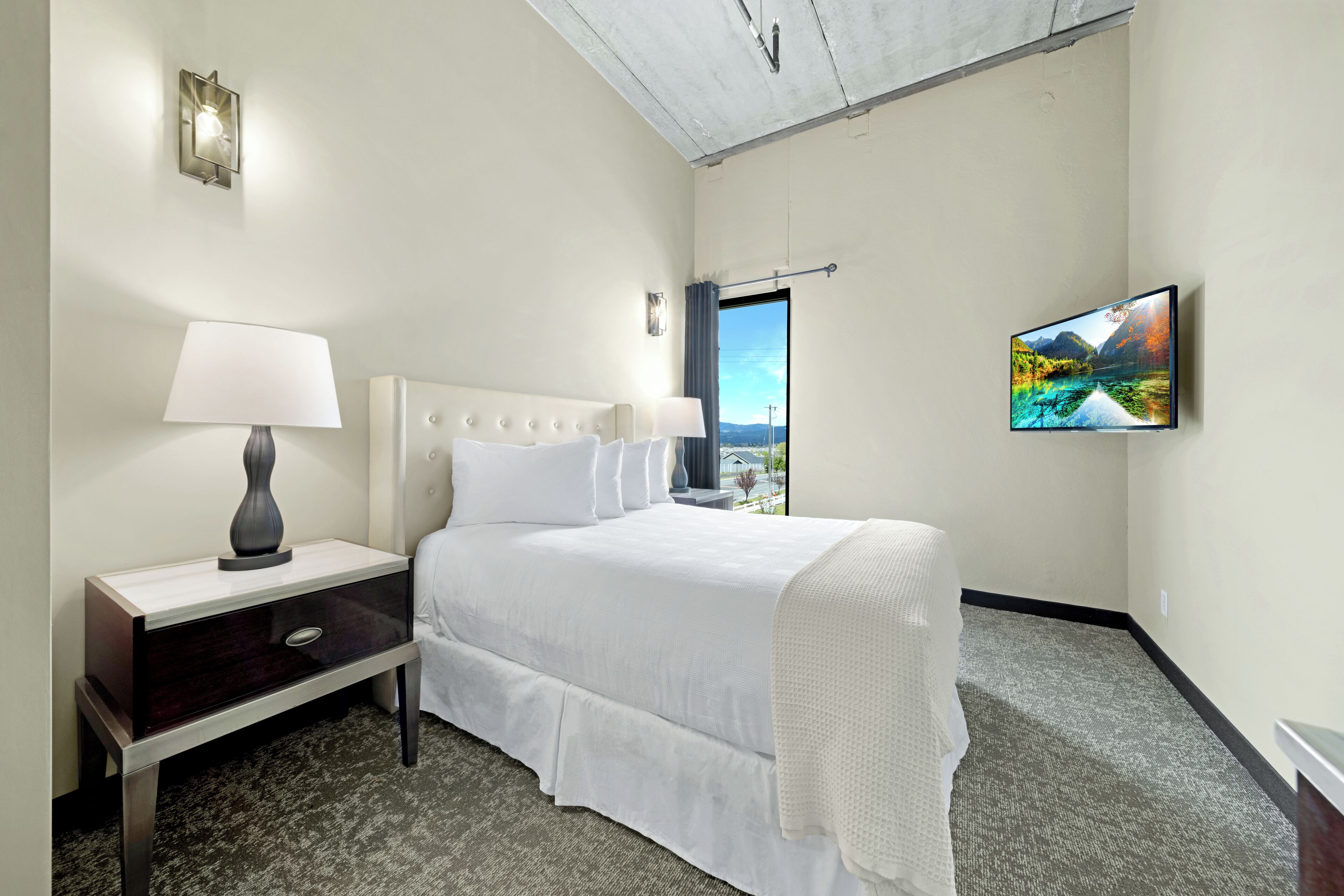 A hotel room with a bed, a nightstand, a lamp, a TV, and a view of the outdoors.
