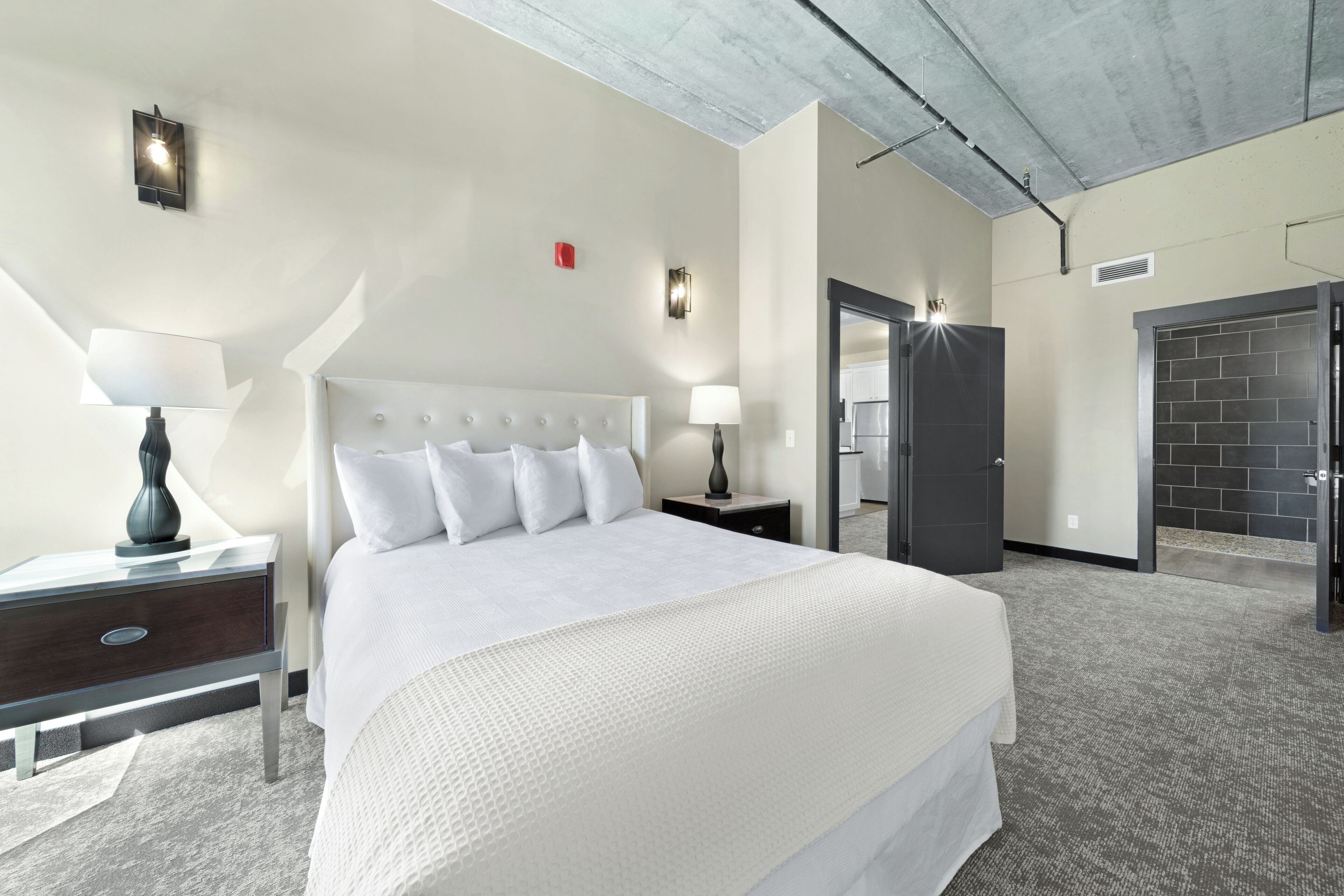 A hotel room with a large bed, two bedside tables with lamps, a modern ceiling, and a door leading to a bathroom.