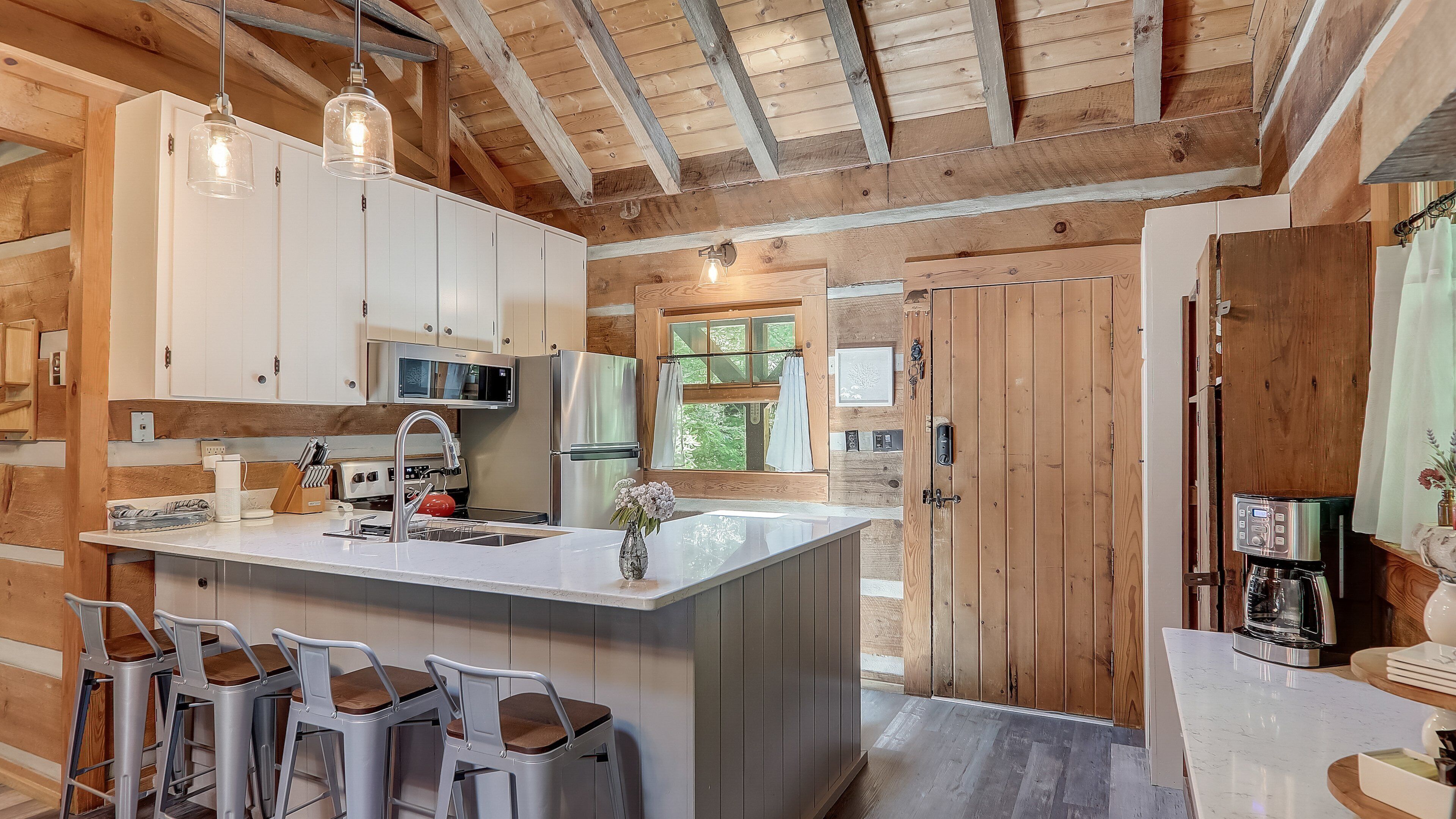 A kitchen with white cabinets, a stainless steel refrigerator, a microwave, and a coffee machine. There are bar stools and a large island with a sink.