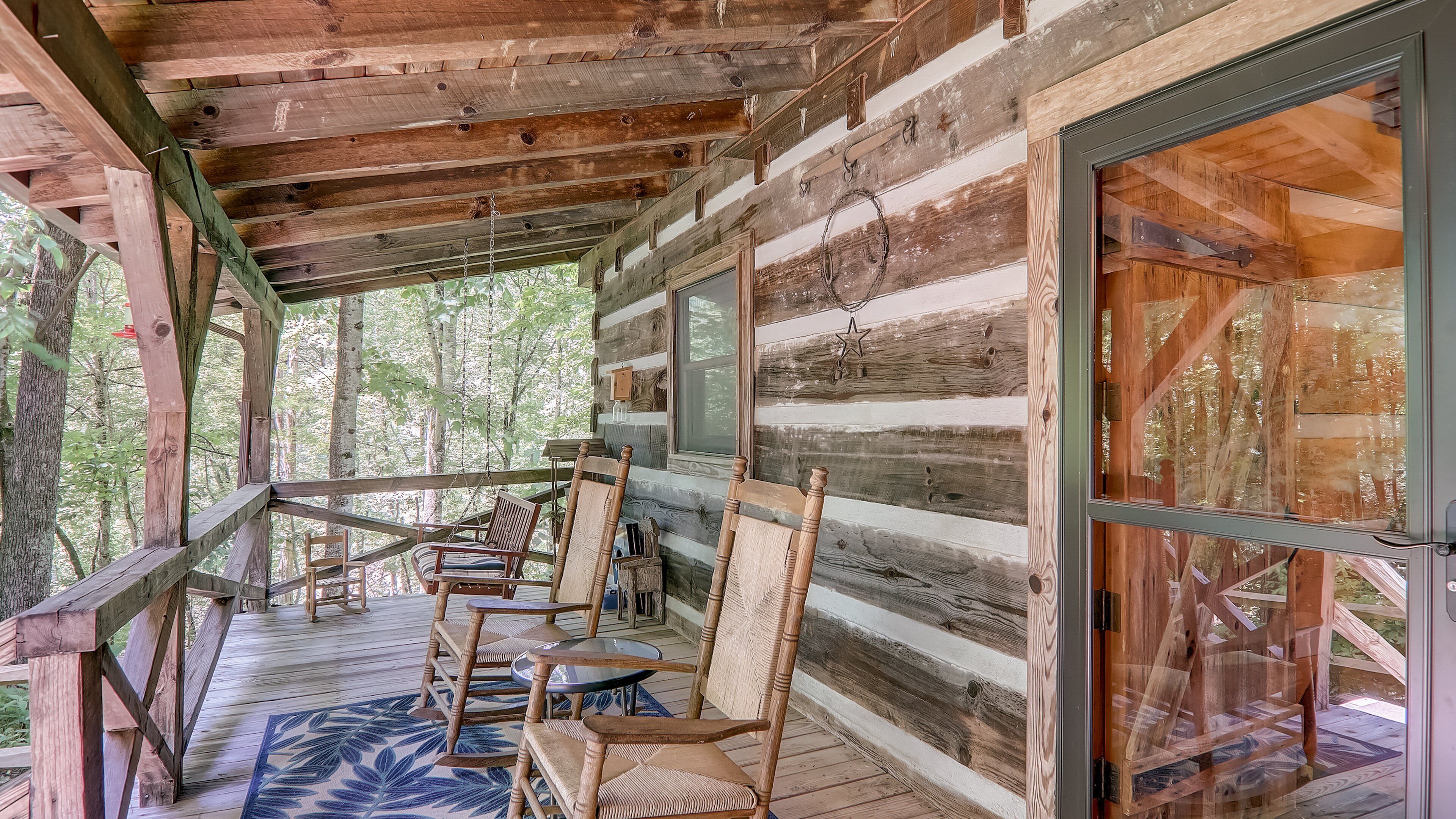 A wooden cabin with a porch, furnished with chairs and a table, overlooking a forested area.