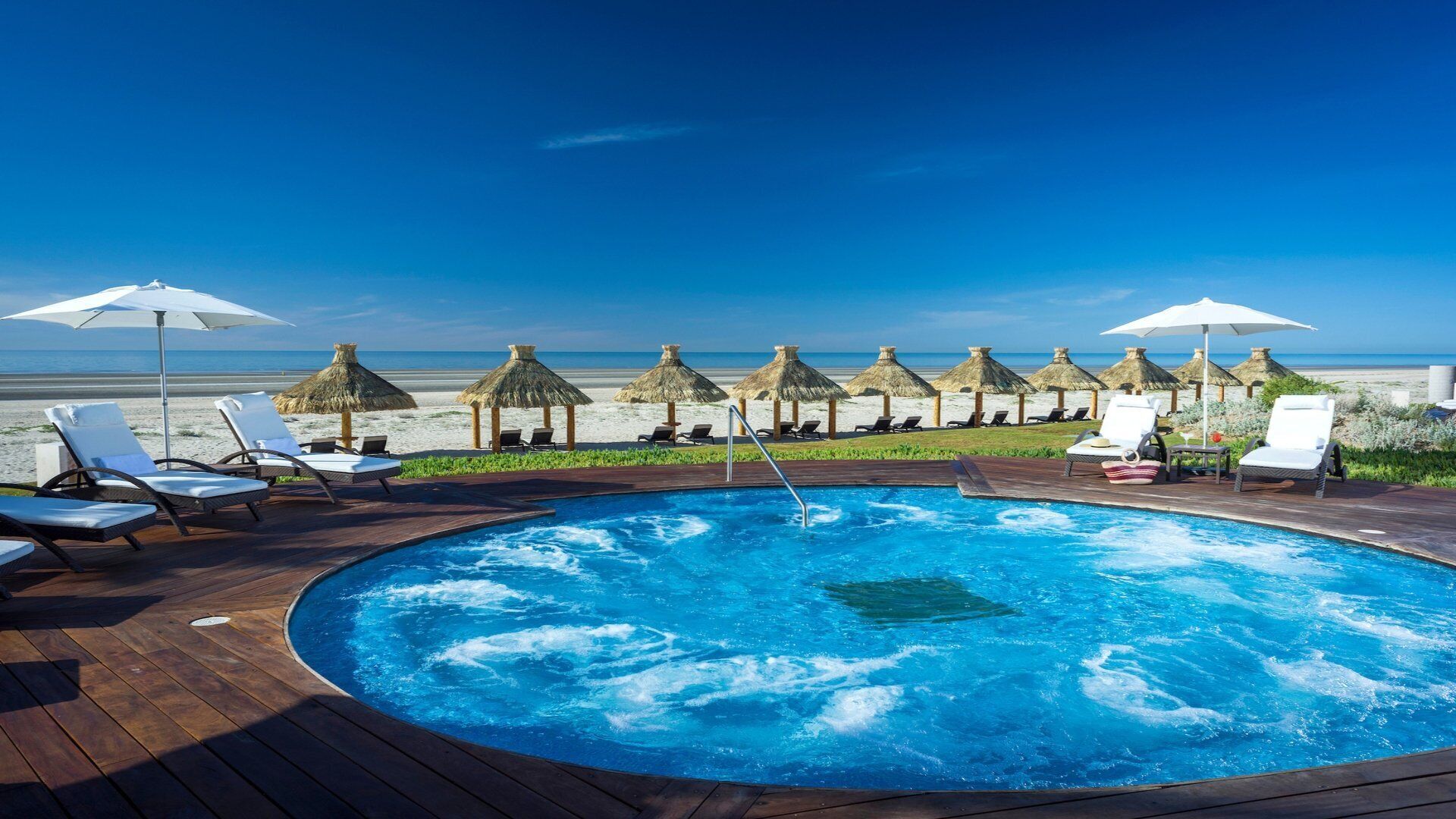 Un espace piscine doté de chaises longues et de parasols, avec vue sur une plage bordée de parasols en chaume.