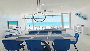 Una sala de estar moderna con una mesa de comedor grande, sillas azules y vista al océano a través de amplios ventanales.