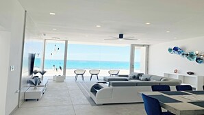 Una sala de estar moderna con un sofá grande, una mesa de comedor y sillas. Se ve el océano a través de ventanas amplias.