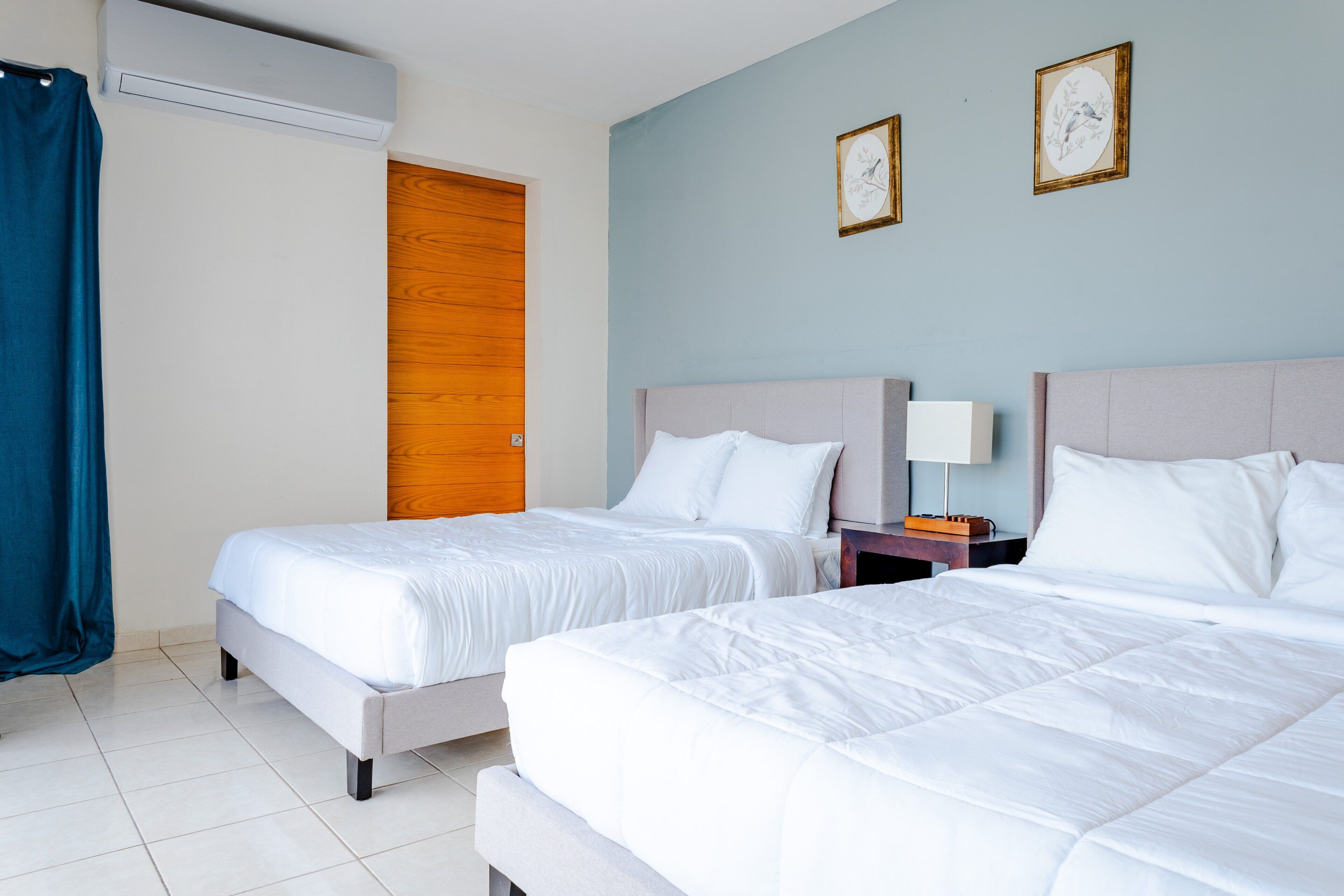 Une chambre d’hôtel avec deux lits, une porte en bois, un rideau bleu et deux tableaux encadrés accrochés au mur.
