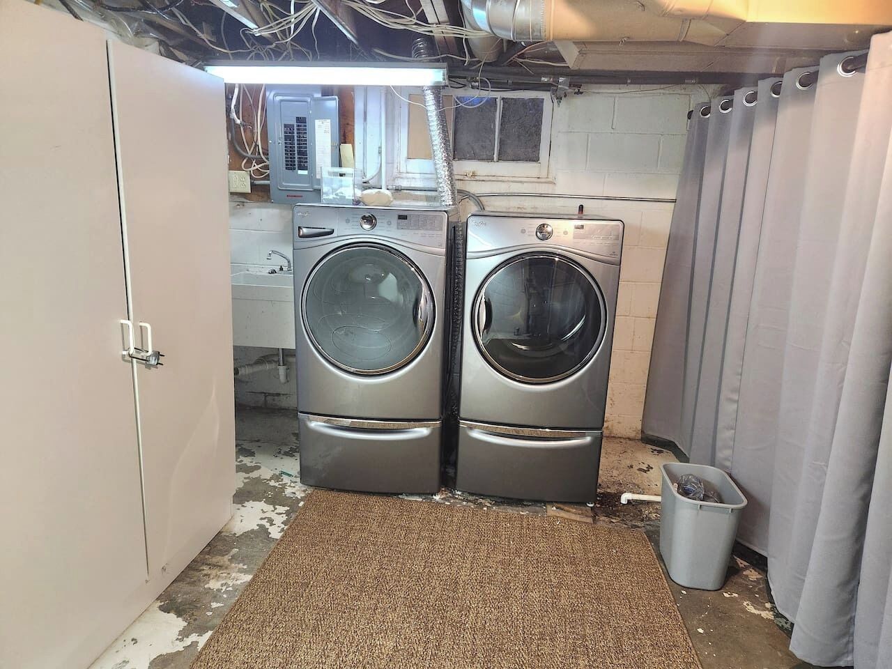 Une buanderie équipée de deux machines à laver à chargement frontal, d’une poubelle et d’un rideau.