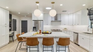 A modern kitchen with white cabinetry, a central island, and four brown chairs. - White Agate by Avantstay Pool + Spa + Putting Green Playground + Outdoor Dining Minutes to Kierland (Scottsdale)