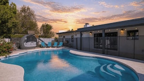 A swimming pool with a curved edge, a covered seating area, and a building with large windows and outdoor lighting. - White Agate by Avantstay Pool + Spa + Putting Green Playground + Outdoor Dining Minutes to Kierland (Scottsdale)