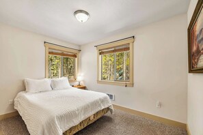 Une chambre à coucher avec un lit, deux fenêtres équipées de stores, un luminaire au plafond et un tableau accroché au mur.
