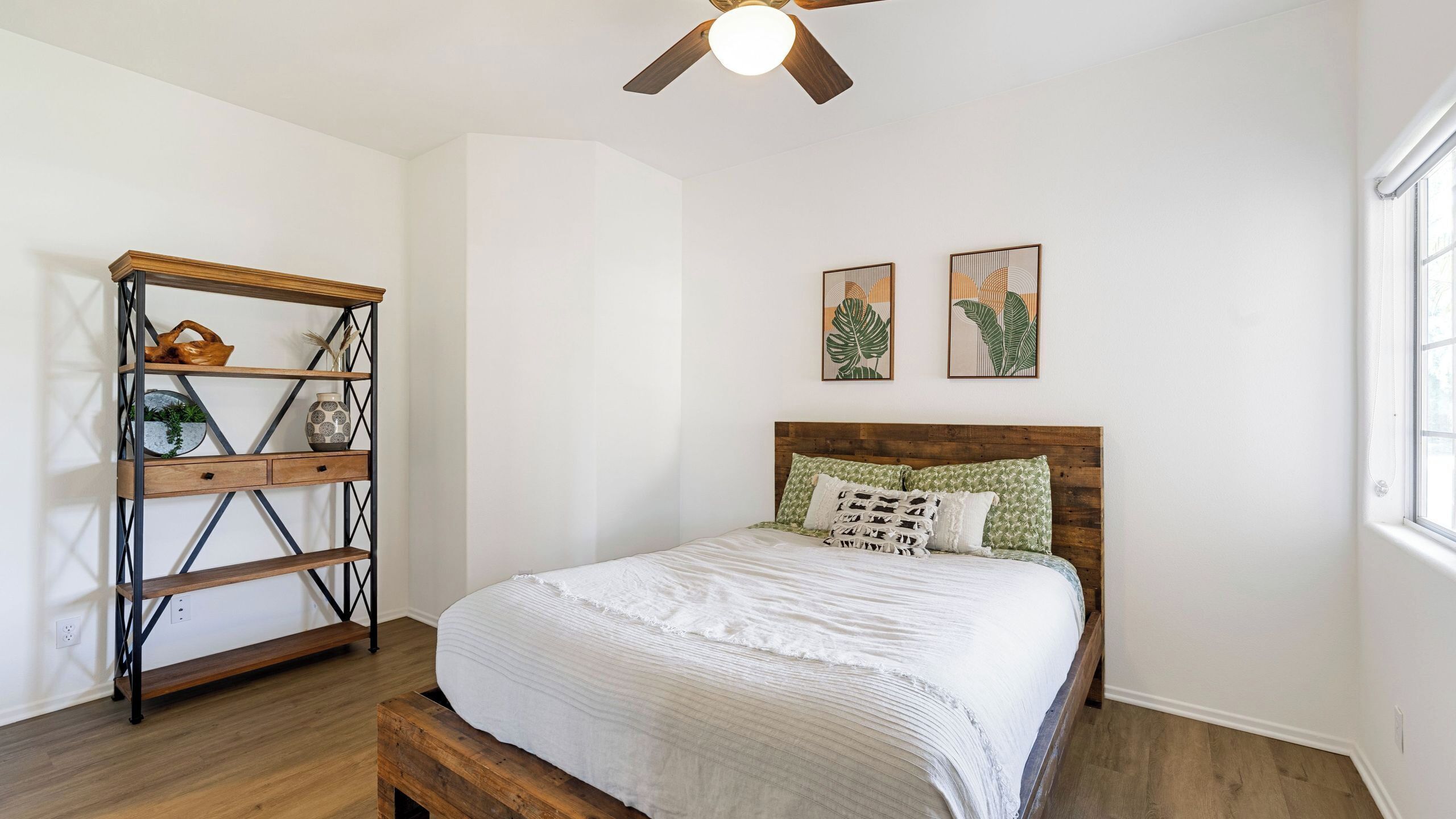 A bedroom with a wooden bed, a wooden nightstand, a metal shelving unit, and two wall-mounted plant prints.