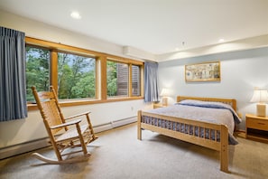Quarto com cama de madeira, uma poltrona de balanço, uma janela grande com cortinas, uma mesa de cabeceira com abajur e um quadro na parede.