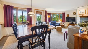 A spacious living room with a dining area, large windows, a television, and a clock on the wall. - 2 bedroom Ski-in condo with pool, hot tub, and EV chargers. (Killington)