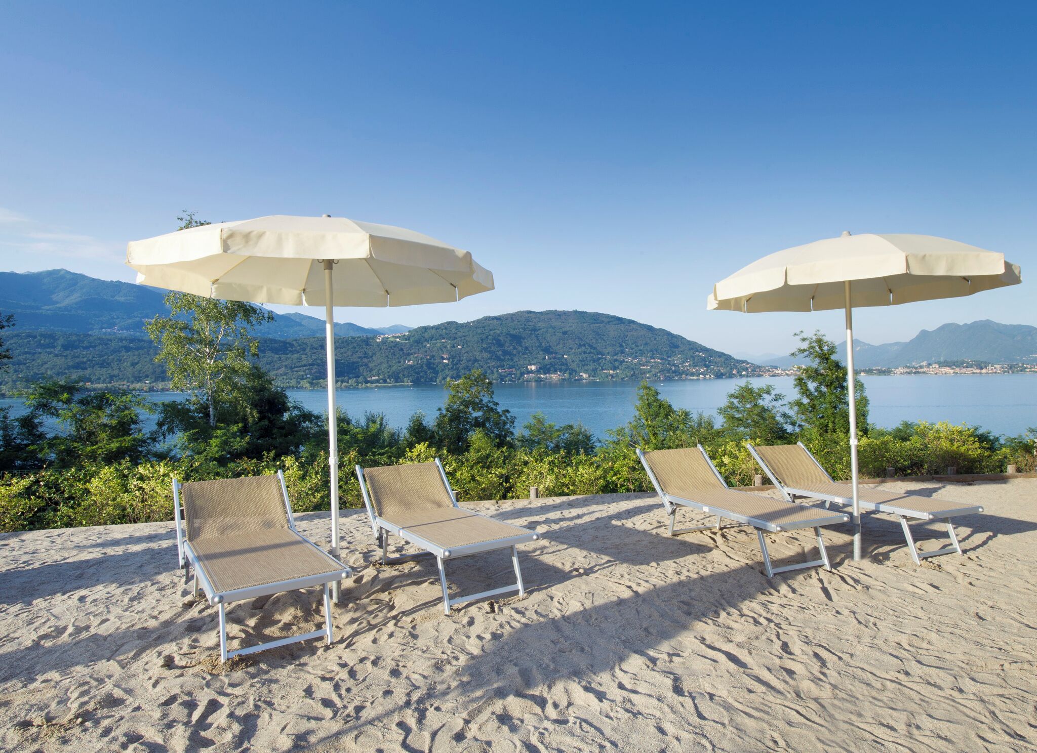 Een strand met vier ligbedden en twee parasols, met uitzicht op een meer en bergen.