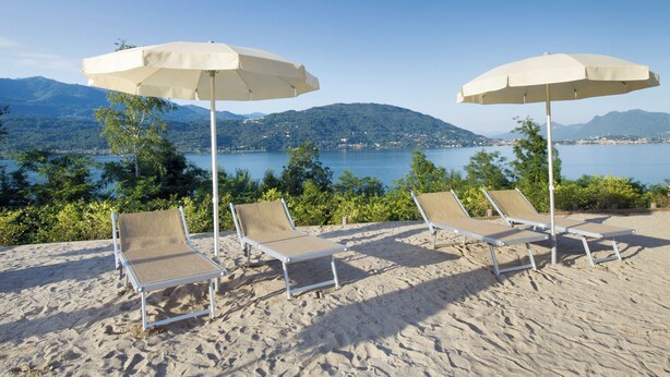 Ein Strand mit vier Liegestühlen und zwei Sonnenschirmen, mit Blick auf einen See und Berge.