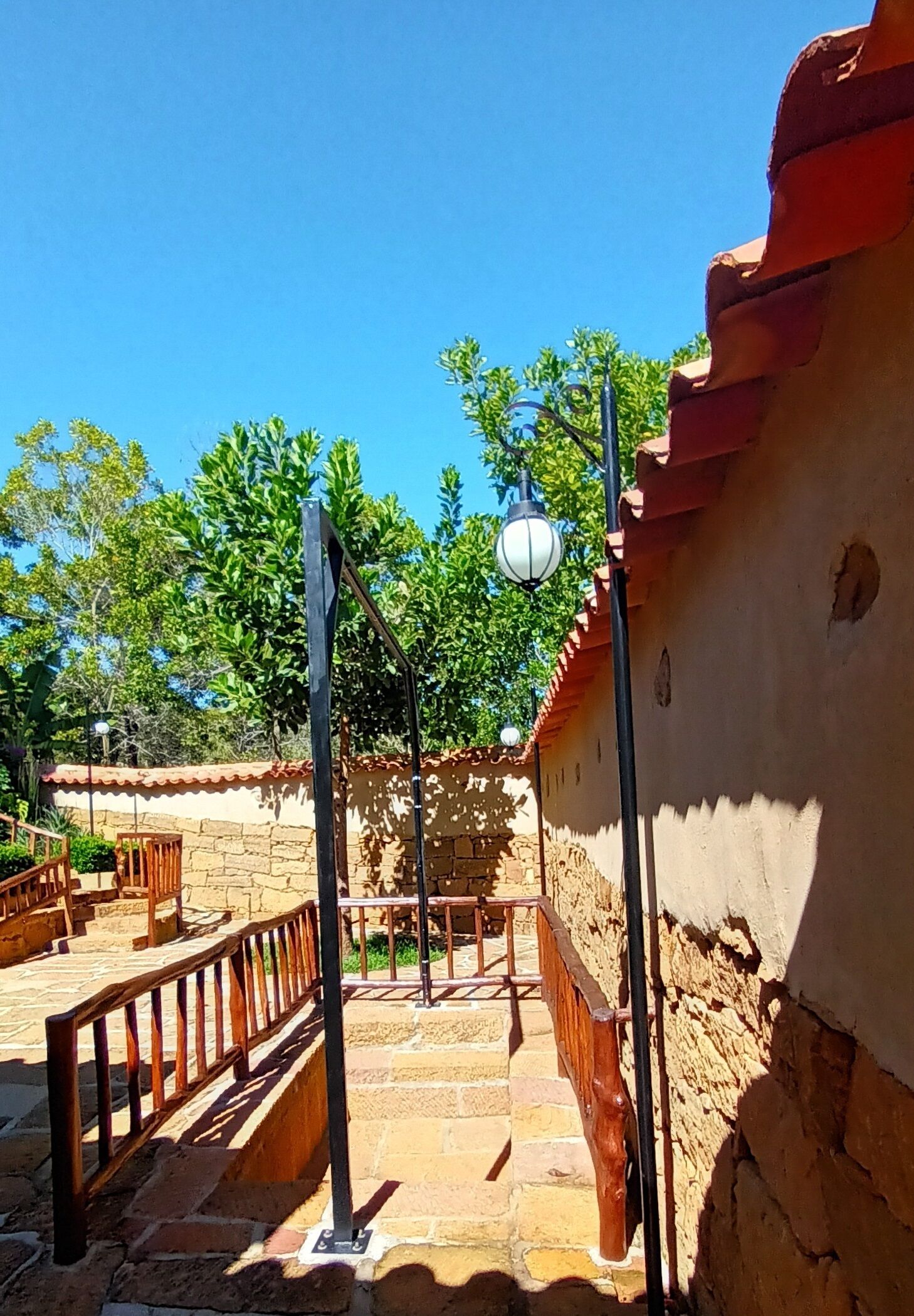 Una escalera de piedra conduce a una terraza de madera con barandilla, rodeada por un muro de piedra y un fondo de árboles.