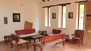 Una habitación con un banco de madera, dos sillas, una mesita y un sofá. Tres ventanas permiten la entrada de luz natural.