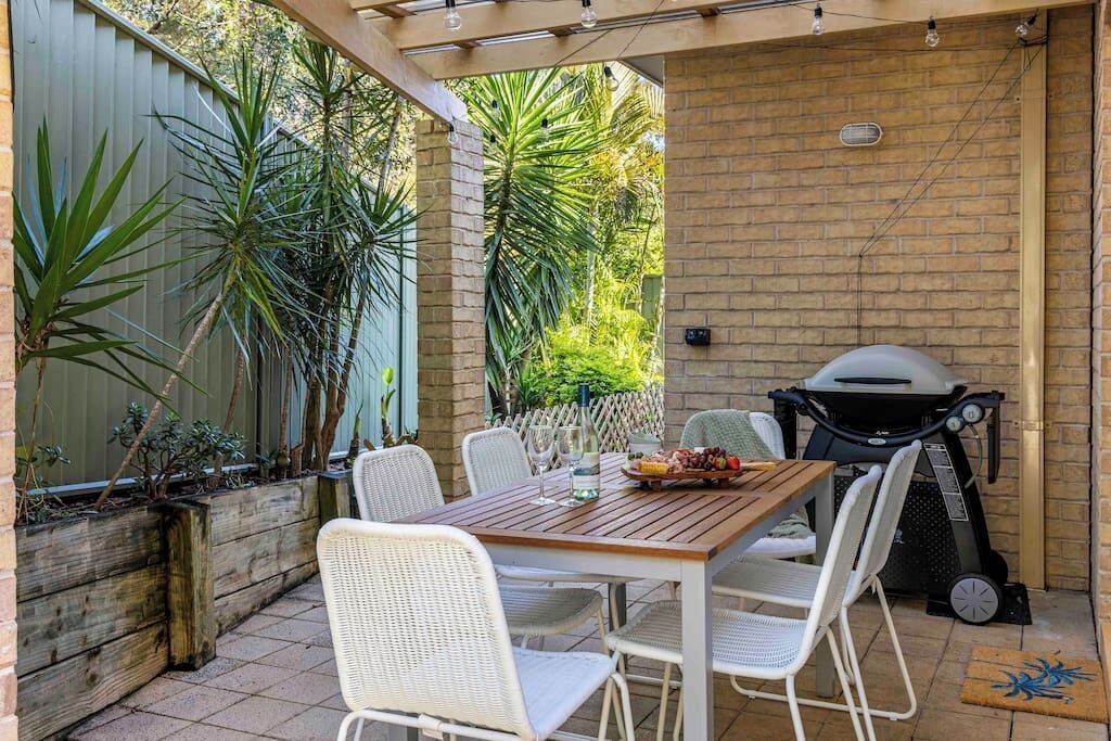 Eine Terrasse im Freien mit Holztisch, weißen Stühlen, einem Grill und Topfpflanzen.
