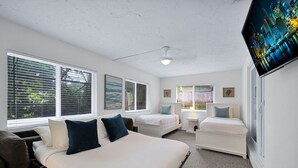 A bedroom with a large bed, two smaller beds, a TV on the wall, and large windows with blinds.