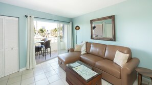 A living room with a brown sofa, a glass coffee table with a map, a dining area with chairs, and a sliding glass door leading to an outdoor balcony.