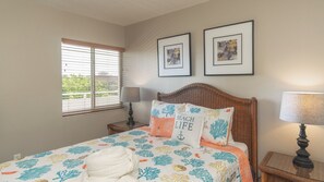 Un dormitorio con una cama, un ventanal con persianas, una mesita de noche con lámpara y dos cuadros enmarcados en la pared.