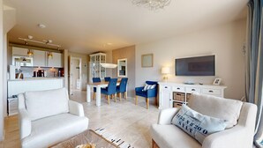 Ein modernes Wohnzimmer mit einer Couch, einem Couchtisch und einem Fernseher. Es gibt einen Essbereich mit Tisch und Stühlen. Im Hintergrund ist eine Küche zu sehen.