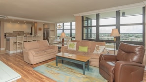 Amplia sala de estar con un sofá beige, un sillón de cuero marrón y una mesa de centro. Hay una zona de cocina con armarios blancos y una mesa de comedor con sillas. Grandes ventanales ofrecen vistas de la ciudad.