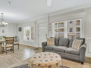 A living room with a gray sofa, a round ottoman with a nautical pattern, a wooden dining table with chairs, and a chandelier.