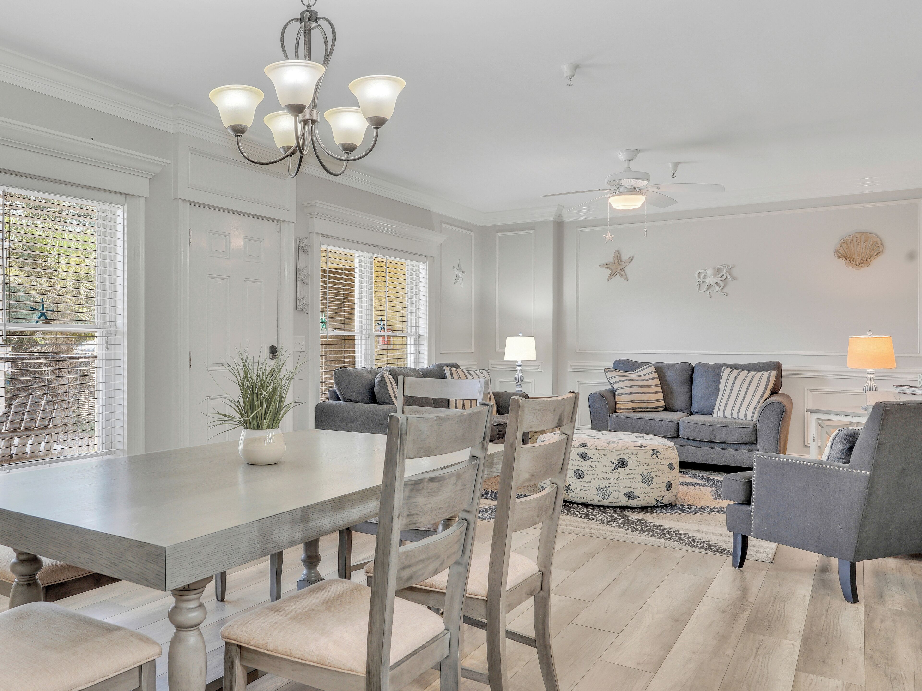 A spacious living room with a dining area, featuring a chandelier, ceiling fan, and decorative wall elements.