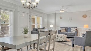 A spacious living room with a dining area, featuring a chandelier, ceiling fan, and decorative wall elements. - Walking Distance to Beach, Heated Pool Access (Tybee Island)