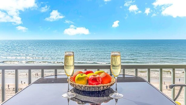 Ein Tisch mit Obstschale und zwei Sektgläsern, mit Blick auf einen Strand und den Ozean.
