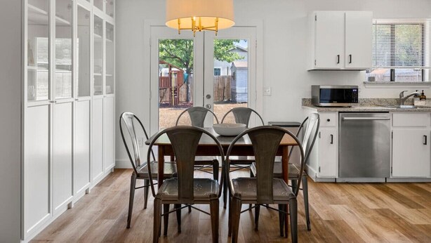 A modern kitchen with white cabinets, a dining table with chairs, a hanging light fixture, and a view through the glass door. - Office, Patio, Yard, Pet Friendly, Playground (Austin)