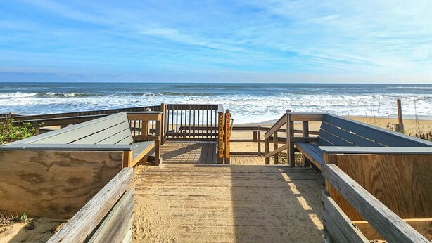 Un paseo marítimo de madera que conduce a una playa, con un banco y un cartel que indica una corta distancia.