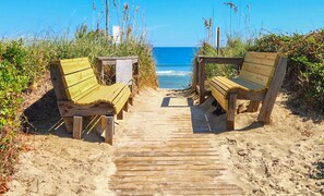 A wooden pathway leading to two wooden benches on a sandy beach with a clear view of the ocean.