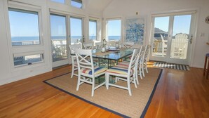 Comedor con mesa de vidrio y sillas blancas, ventanas amplias con vistas al océano y piso de madera.