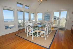 A dining area with a glass table and white chairs, large windows with ocean views, and a wooden floor. - 5th Ave Coastal Retreat - 5 BR Oceanfront home w/Private Pool! (Southern Shores)