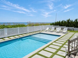 Une piscine rectangulaire à l’eau d’un bleu cristallin, entourée d’une clôture blanche et d’une terrasse en bois agrémentée de chaises longues blanches.